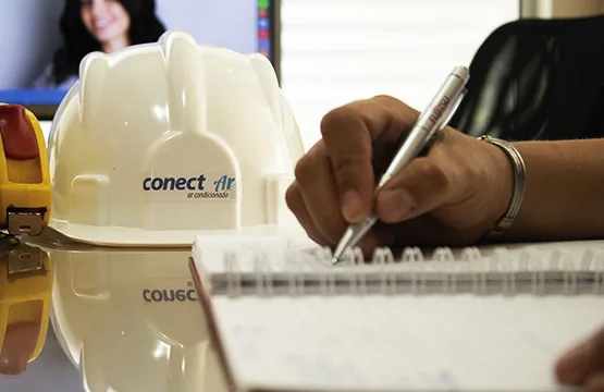 Capacete branco com o logotipo da Conect Ar sobre a mesa, ao lado de mãos anotando medidas em um caderno